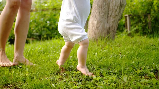 Little cute toddler boy holding his mother's hands and doing first steps barefeet on the grass.