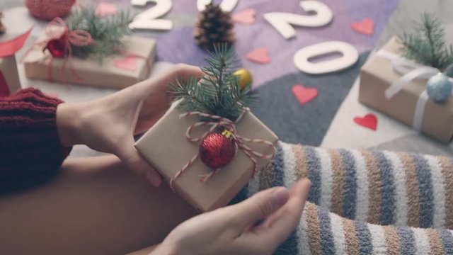 Young woman hands making red and white twine bow on craft paper gift box on her knees in cozy home indoor. Young girl preparing for festive holiday season.