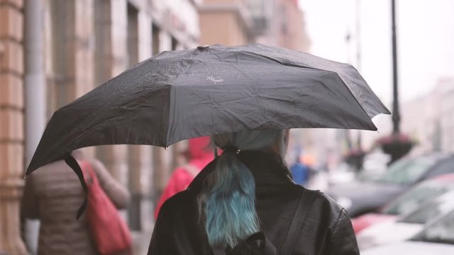 Woman with black umbrella walking on the street under the rain. Buildings and cars on background. Slow motion.
