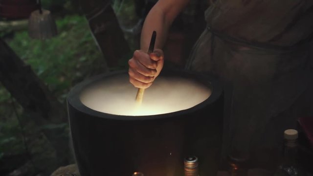 human hand of old woman evil witch is stirring potion in  bubbling vat pot with wooden spoon. mystery silhouette preparing cooking potion medicine couples spell, curse. Halloween sorceress mage. 