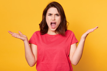 Close up portrait of angry disappointed brunette shouting with dissatisfaction, opening her mouth widely, looking directly at camera, raising her hands, standing isolated over yellow background.