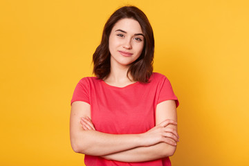 Fototapeta premium Good looking adorable young woman looking directly at camera, having pleasant facial expression, having short black hair, standing with folded arms, wearing casual red t shirt. Emotions concept.