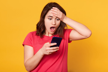 Fototapeta premium Portrait of shocked emotional woman putting one hand to forehead, holding her smartphone, opening her mouth widely with surprise, looking at her mobile phone screen, reading news. Emotions concept.