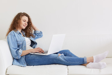 Naklejka premium Horizontal shot of good looking curly haired young model sitting on white sofa, having laptop, watching videos, applying information, wearing jeans, white t shirt, socks and jacket. Tech concept.
