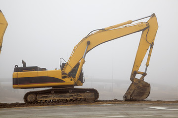 Excavator on road at a construction site with large industrial equipment in the fog
