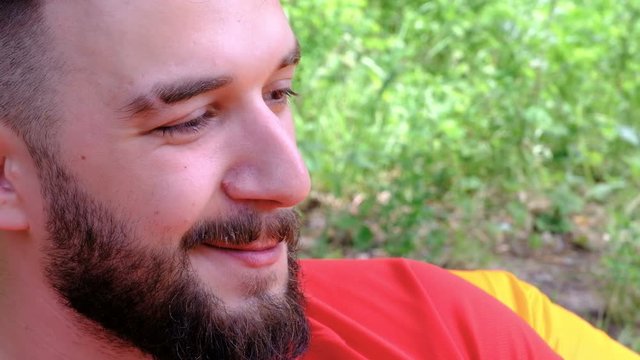 A young man in a red T-shirt drinks beer with pleasure. Close-up. The guy with the beard drank a frothy alcoholic beverage out of a glass. Rest, picnic and travel. Concept