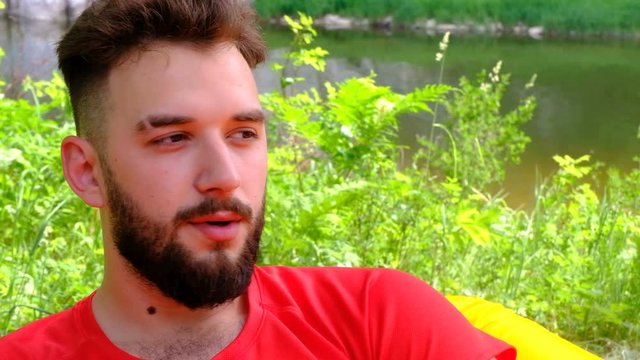 A young man with a beard enjoys drinking coffee in nature. Closeup of a brutal guy with a white cup of tea in his hand against the background of mountains and a river.