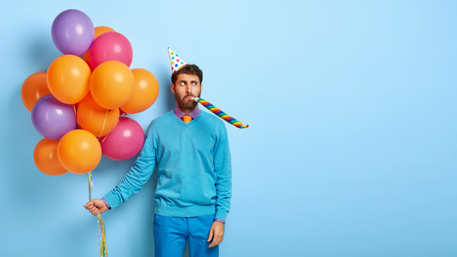 Serious man wears cone birthday hat, blows in party whistle, holds colorful air balloons, dressed in neat blue clothes, stands indoor, celebrates anniversary. Blank copy space for your information