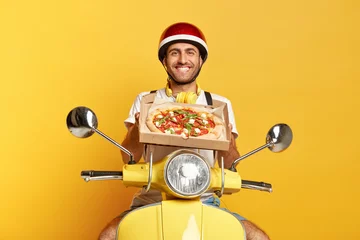 Poster Satisfied motorcyclist delivers tasty fresh baked pizza, wishes good appetite for customers, holds cardboard box, wears protective helmet, sits on fast motorbike, isolated on yellow studio wall © Wayhome Studio