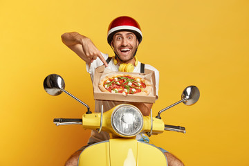 People, food concept. Glad delivery man points at appetizing italian pizza, suggests to taste, conveys for clients, drives fast motorbike, wears protective helmet, poses over yellow background.