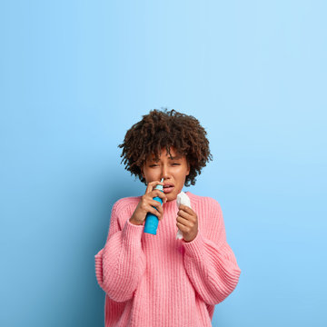 Photo of ill Afro woman sprays drops in nose, holds handkerchief, being allergic to something, sneezes, wears pink casual sweater, isolated on blue wall with blank copy space. People and sickness