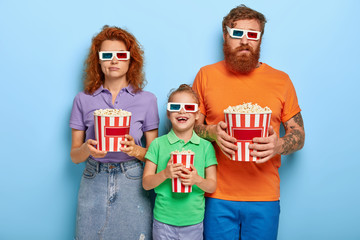 Bored ginger father and mother come on cinema together with small daughter for watching new cartoon in 3d, wears dimension stereo glasses, eat popcorn. Glad child spends time with loving parents