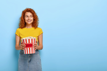 Photo of ginger charming girl holds bucket with popcorn, going to watch movie at home, waits for friend, wears yellow t shirt and denim skirt, relaxes indoor, isolated on blue wall. Spare time concept