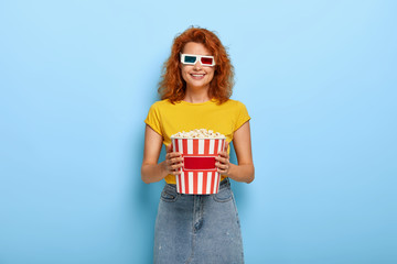 Photo of happy red haired millennial female wears 3d glases, eats tasty popcorn, enjoys watching favourite film, smiles gently, dressed in fashionable outfit. Pretty viewer with junk food in cinema