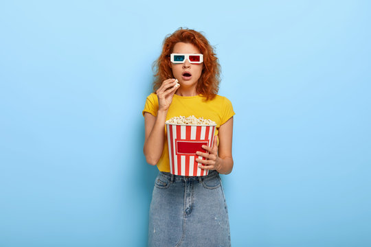 Beautiful shocked woman has impressive look, watches fantastic movie, wears special glasses and visits 3d cinema, eats popcorn, isolated on blue background, cannot miss any second, being frightened
