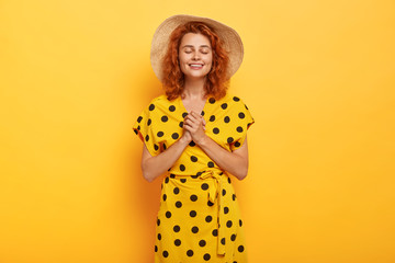 Gentle pleased female keeps hands together, eyes shut, prays for well being of her family, has hope in better, wears stylish polka dot dress, hat stands in studio over yellow background. Body language