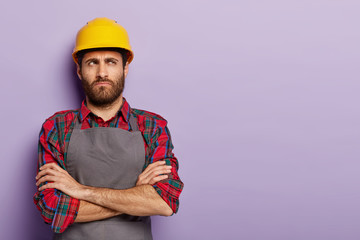 Shot of pensive dissatisfied construction worker in protective yellow helmet, stands with arms...