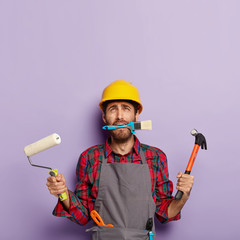 Home repair male worker busy with house renovation, holds building equipment, has sad expression,...