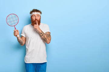 Athlete hipster with tennis racket in hand, makes shush gesture, wears sport clothes, tells secret, looks surprisingly away, has tattoo, stands against blue background, copy space for your promotion