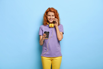 Beautiful glad female has funny conversation, uses modern cell phone, drinks takeaway coffee, has relaxed look, calls friend, has curly red hair, dressed in bright clothing isolated on blue background