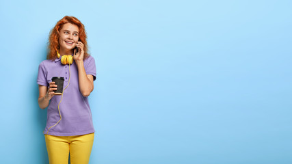 Joyful cute ginger student in trendy outfit, drinks coffee and talks on mobile phone, looks aside with dreamy expression, uses headphones, isolated on blue wall, blank space for your advertisement