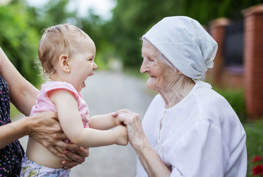 Happy Toddler Girl And Her Great Grandmother Holding Hands And Looking At One Another