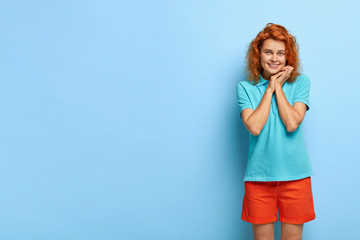 Happy shy girl keeps hands near face, smiles gently at camera, looks with pleasure, dressed...