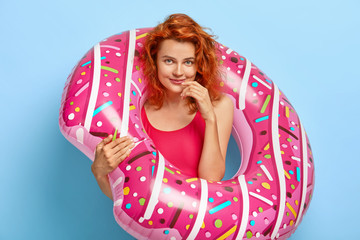 Photo of lovely ginger woman looks happily, poses inside of rubber swim ring, dressed in swimwear, rests at seaside, has blue eyes, healthy skin, models indoor. People, summer vacation concept