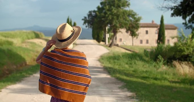 Back view attractive stylish woman holding hat walking on the country road in popular italian valley in Tuscany. Excited female tourist in Val d&rsquo;Orcia region. Tourism. Summer trip.