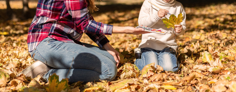 Girl and her mother sitting on a autumn leaves in the park. Drawing with paint autumn leaves outside