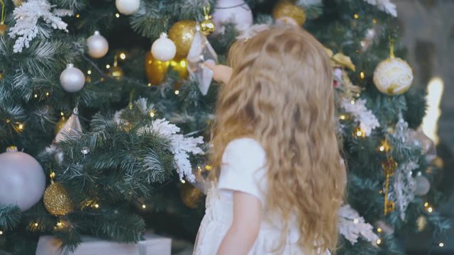 funny little blond girl in elegant dress touches balls on beautiful decorated Christmas tree at home