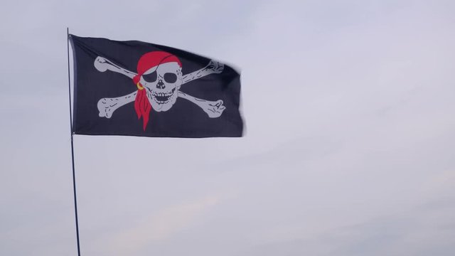 Pirate flag Jolly Roger hanging on a ship mast on a blue sky background at windy day.