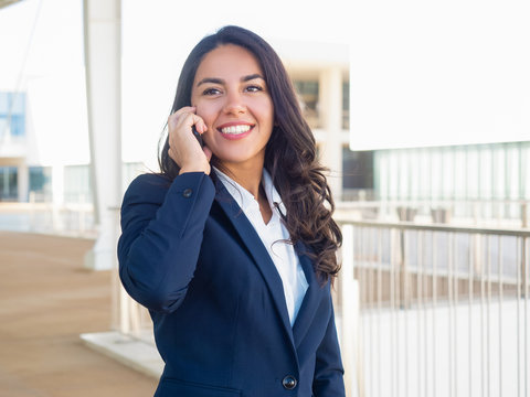 Happy confident professional talking on cellphone outside. Beautiful young Latin woman in office suit calling on phone during work break. Business phone talk concept