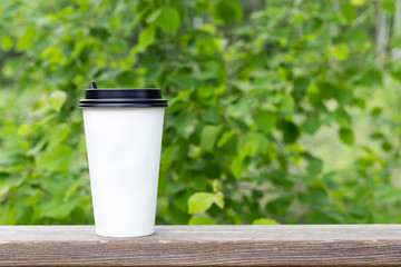 White paper cup with black lid on wooden stand, green summer park background. Take away, coffee shop concept. Close-up, copy space, mock up