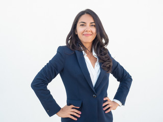 Proud confident business woman posing over white background. Happy beautiful young Latin woman in office suit placing hands on hip, looking at camera and smiling. Business success concept
