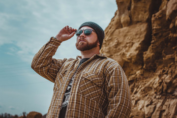 brutal man in glasses and a hat on the background of rocks