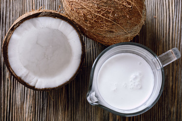 Thick coconut milk in a glass milk jug next to half a opened coconut and a whole coconut on a wooden table, close-up, top view