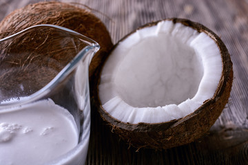 Thick coconut milk in a glass milk jug next to half a opened coconut and a whole coconut on a wooden table, close-up