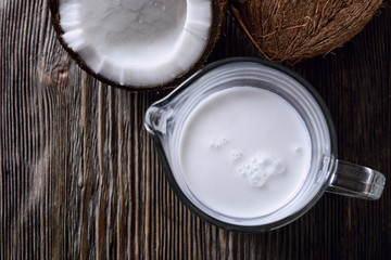 Thick coconut milk in a glass milk jug next to half a opened coconut and a whole coconut on a wooden table, close-up, top view