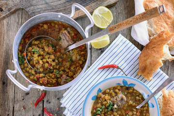 Mixed legumes and peas soup with meat in a vintage aluminum pan with a ladle for serving on an old wooden table next to a bowl with this soup, slices of tortilla, lime and chili pepper.