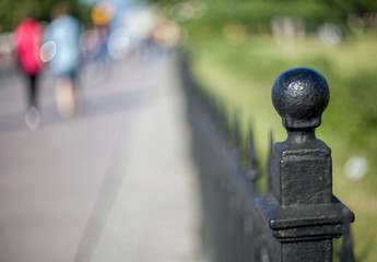 Metal pommel in the form of a ball on the cast-iron fence of artistic casting in a park in St. Petersburg with a blurred background of people walking along the path.