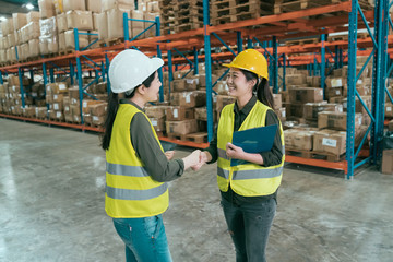 Warehouse operator teamwork. two female storehouse owner partners shaking hand. young asian woman in hard hats handshaking and smiling with great deal while standing in large stockroom together.