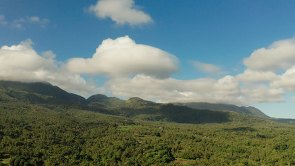Mountains covered rainforest, trees and blue sky with clouds, aerial view. Camiguin, Philippines. Mountain landscape on tropical island with mountain peaks covered with forest. Slopes of mountains