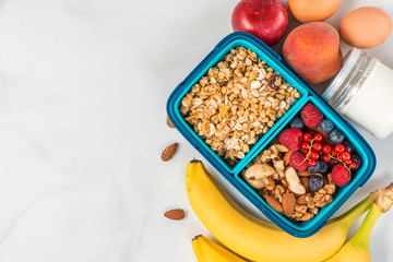 school lunch box with granola, nuts, berries, banana, eggs, peaches and yogurt to make breakfast on white background