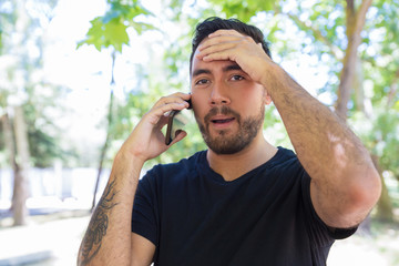 Emotional man talking by smartphone and looking at camera. Shocked young bearded man talking by cell phone outdoor. Communication concept