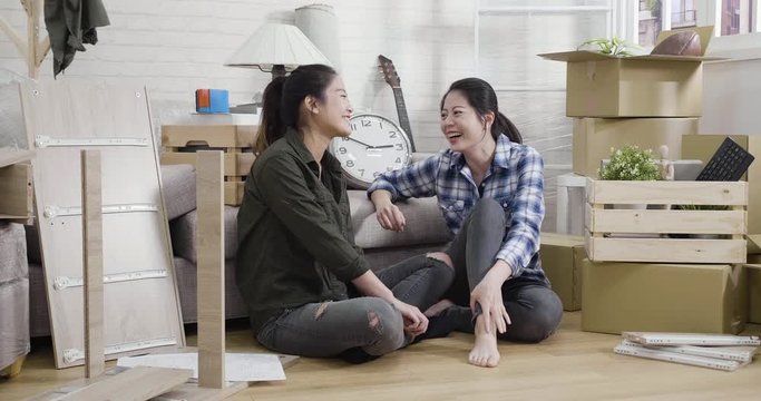 Young family asian sisters relax while assembling furniture at new house. two girls roommates sitting in living room new apartment chatting on floor laughing cheerful surrounding by packing boxes.