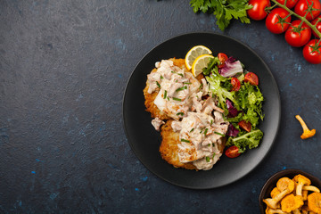 Fried cod, served on potato pancakes with mushrooms sauce and salads. Portion on a black plate. Dark background.