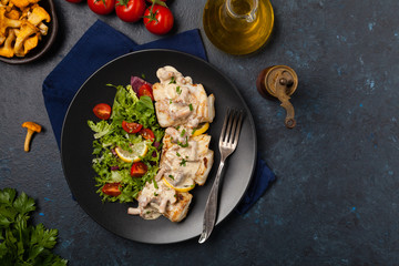 Fried cod served with mushroom sauce and salads. Portion on a black plate. Dark background.