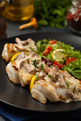 Fried cod served with mushroom sauce and salads. Portion on a black plate. Dark background.