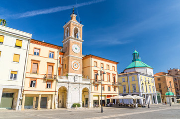 Piazza Tre Martiri Three Martyrs square with traditional buildings with clock and bell tower in old historical touristic city centre Rimini with blue sky background, Emilia-Romagna, Italy - obrazy, fototapety, plakaty